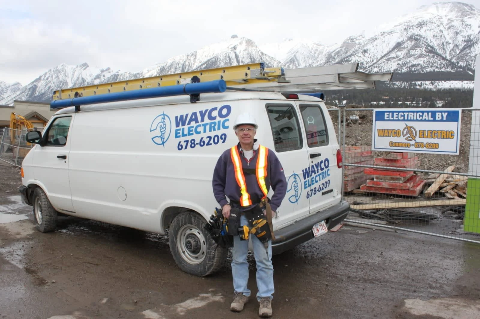 Wayne Cote of Wayco Electric standing beside the company van at a Canmore job site, snow-capped Rockies behind